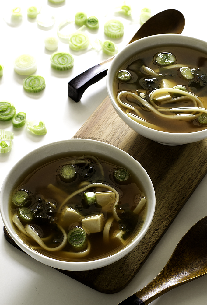 Two small white bowls with a dark brown soup, seaweed pieces, tofu cubes, noodles and chopped scallion.