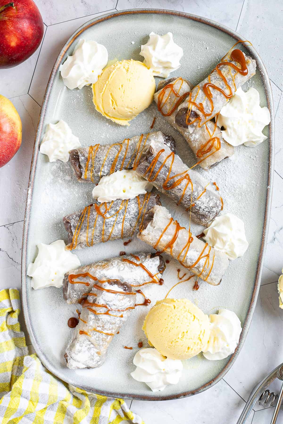 Rice paper rolls with brown filling placed on a light blue plate. Dusted with powdered sugar. Served with scoops of vanilla ice cream, caramel drizzle, and heaps of whipped cream.