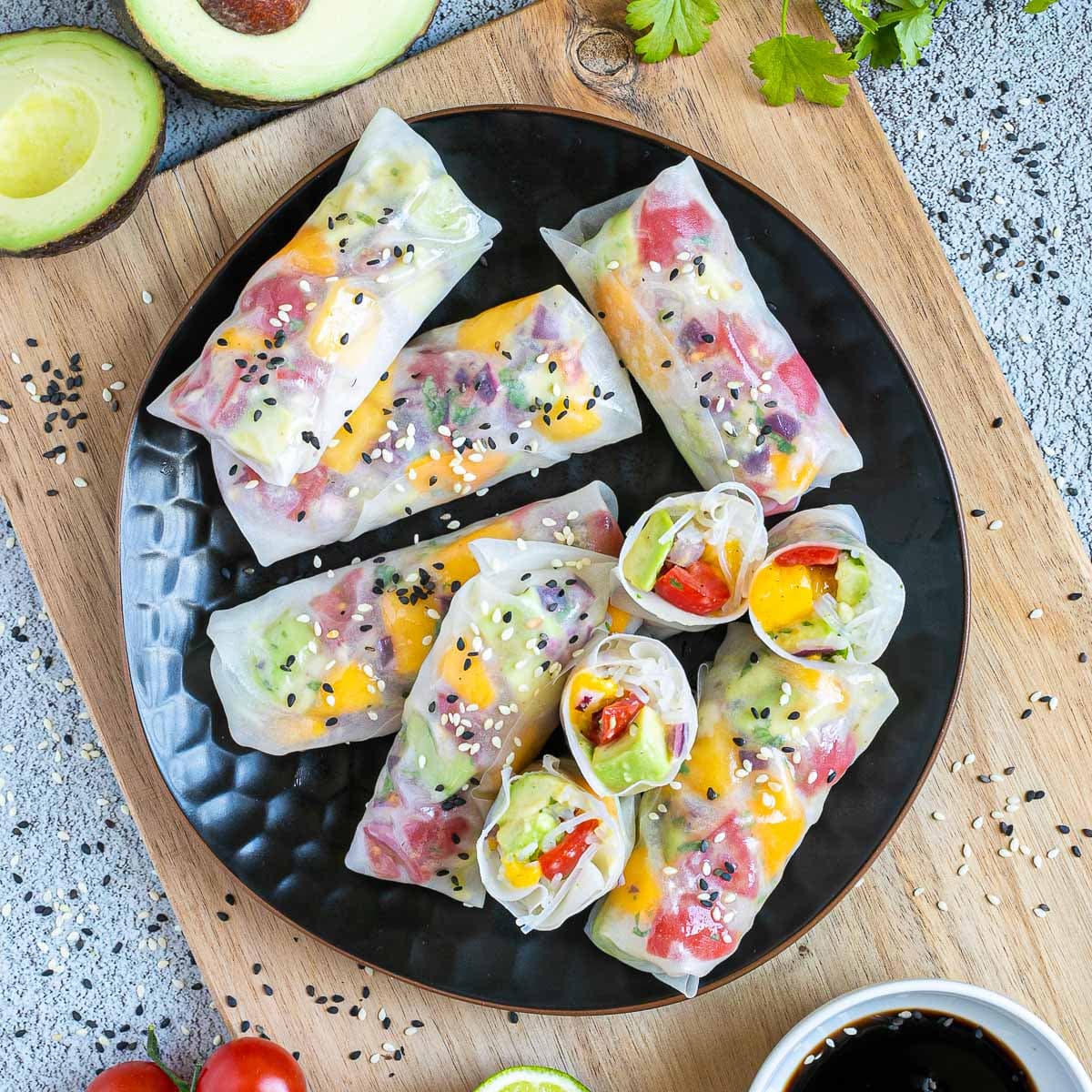 Fresh rice paper wraps filled with chopped tomato, mango, and avocado placed on a black plate. 4 half rolls and 6 whole rolls. Leftover ingredients are next to it.