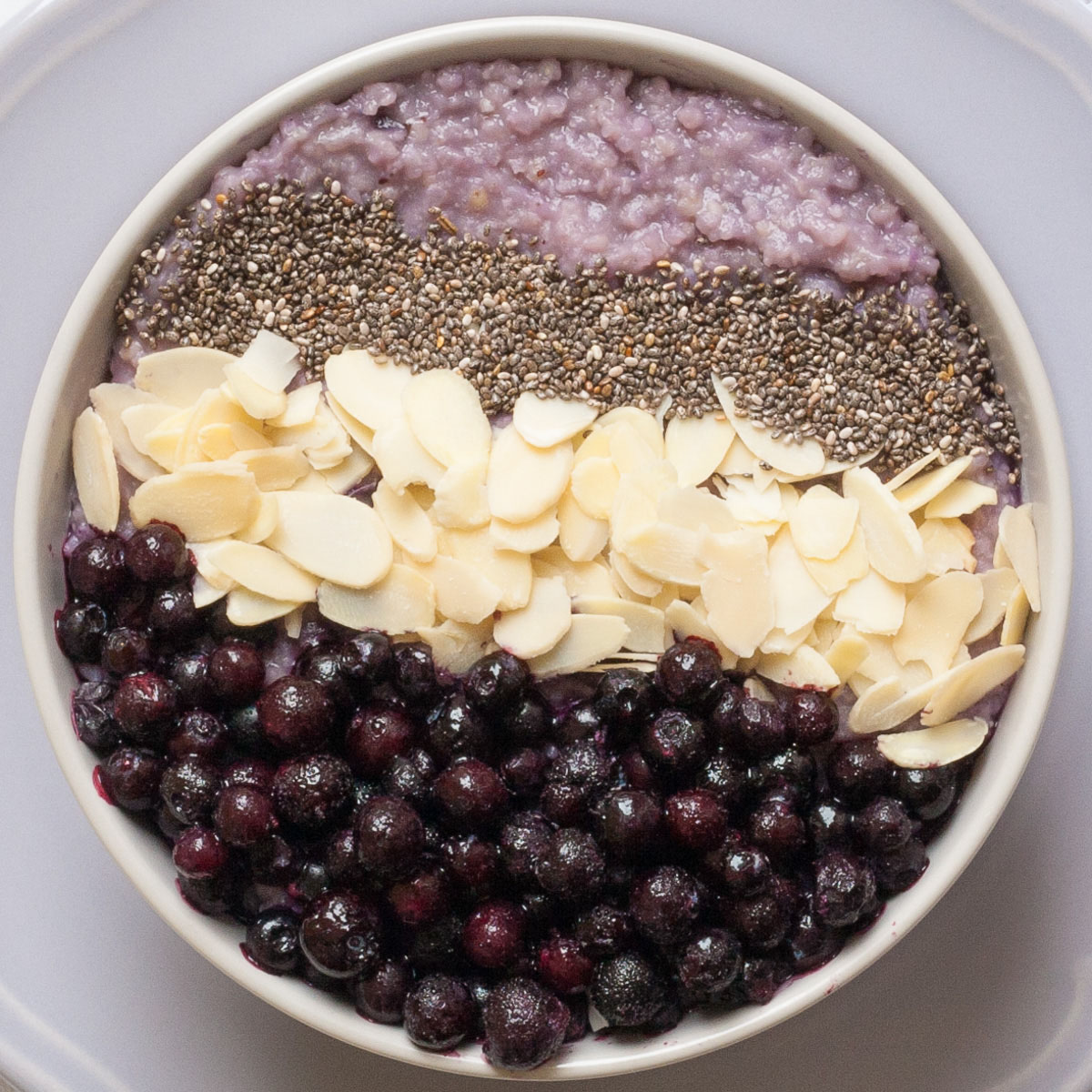 A white bowl on a light purple plate full of purple porridge topped with small grey seeds, almond slices and lots of purple blueberries
