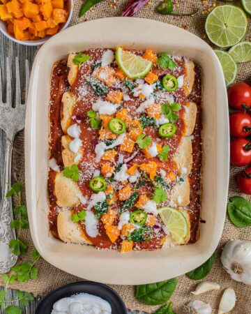 A casserole pan with rolled tortillas covered in red sauce and topped with diced roasted butternut squash, chili pepper slices, cilantro leaves, lime slices, sour cream and grated cheese. More ingredients in small bowls right next to it.