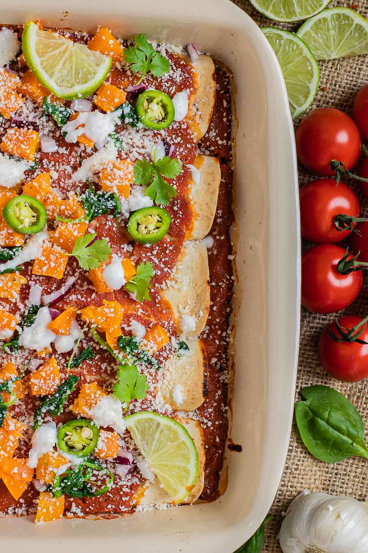A casserole pan with rolled tortillas covered in red sauce and topped with diced roasted butternut squash, chili pepper slices, cilantro leaves, lime slices, sour cream and grated cheese. Tomatoes, lime, garlic, and spinach leaves are right next to it.