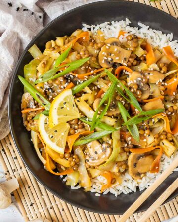 Black plate with white rice topped with stir fired cabbage, mushroom, carrot and lentils. A pair of chopsticks is placed on the side.