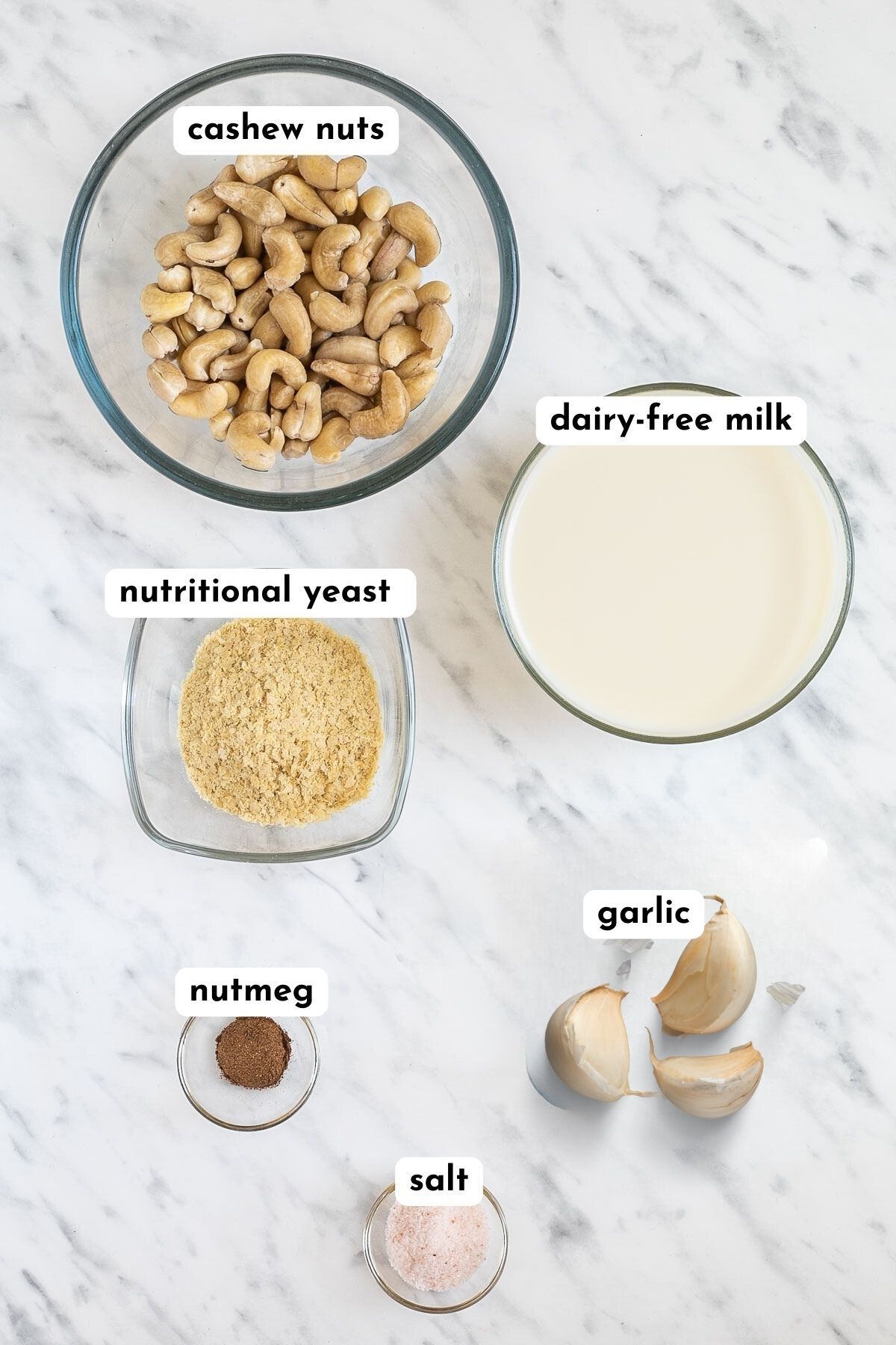 The ingredients of cashew alfredo sauce is in small glass bowls like cashew nuts, milk, yellow flakes, garlic cloves, brown spice and salt
