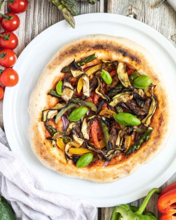 Cheeseless pizza on a white pizza plate topped with tomato sauce, bell pepper strips, zucchini, mushrooms, asparagus, red onion, and basil. More fresh ingredients are next to it.