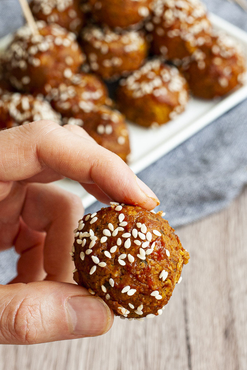 A hand is holding a brown-red ball sprinkled with sesame seeds. In the background more balls on a white plate.