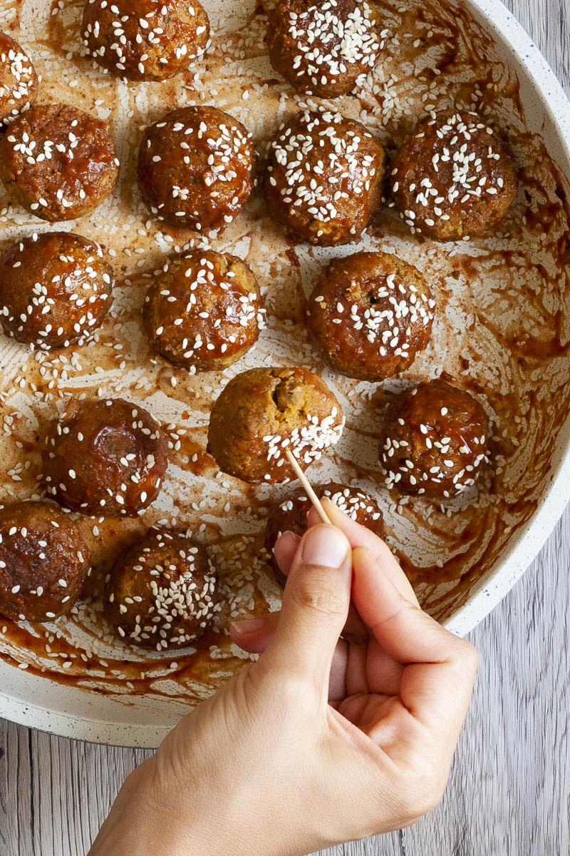 Frying pan from above with several brown-red balls sprinkled with sesame seeds. A hand holding a toothpick takes one ball.