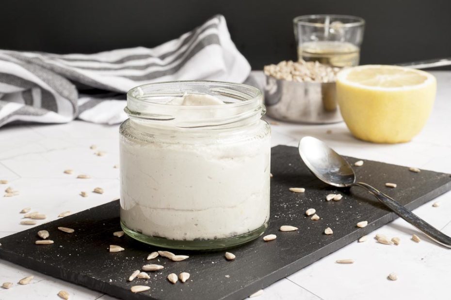 A small glass jar with white paste. Sunflower seeds are scattered around it on a black board. 