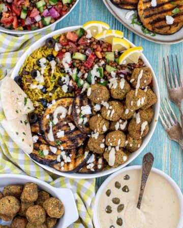 A large white bowl with falafel balls, grilled eggplant, yellow rice and chopped tomato and cucumber salad with lentils drizzled with a white sauce. The remaining ingredients are in bowls around the main one.