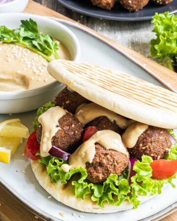 Two pita breads stuffed with 4 dark brown falafel balls, drizzled with a light brown sauce with sliced purple onion, tomato pieces and lettuce. All elements of the sandwich is around it on different plates.
