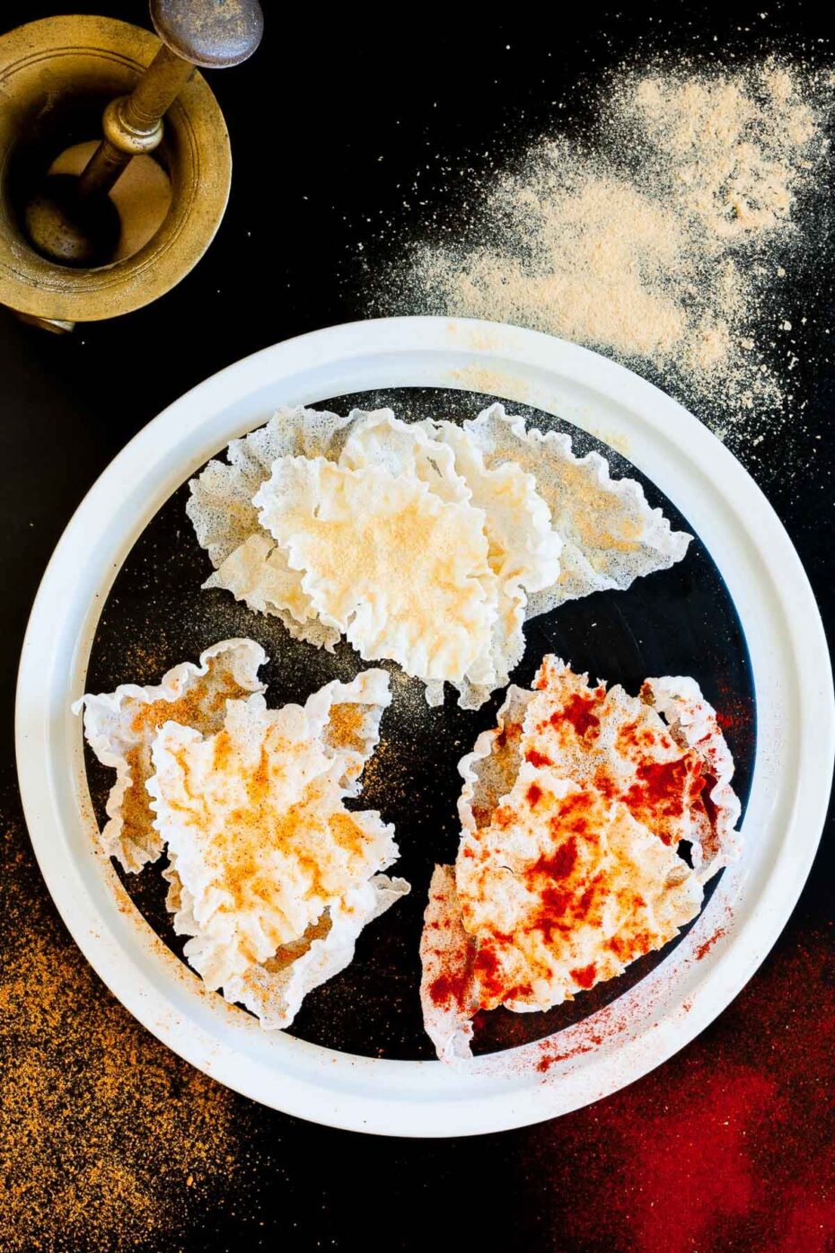 Several puffed rice paper sheets sprinkled with different spices red yellow and brownish on a black plate and those spices are also sprinkled in the black board for contrast