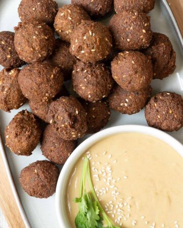 Falafel balls are served on a large plate. A light brown sauce in a white shallow bowl sprinkled with white sesame seeds and topped with a twig of parsley is next to them..