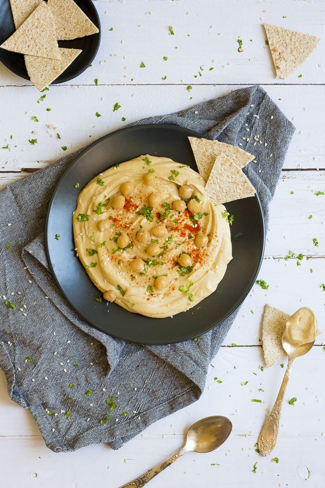 The smoothest, vegan, gluten-free hummus that you can imagine. You only need basic, whole food ingredients AND FIVE MINUTES to make the best oil-free hummus ever. Once, you taste it, you will make it over and over... #glutenfreehummus #oilfreehummus #veganhummus #vitamixhummus #homemadehummus Black plate on grey tablecloth with oil-free hummus spread in the middle sprinkled with cooked chickpeas, fresh parsley, sweet paprika and sesame seeds. Flatbread triangles scattered around.