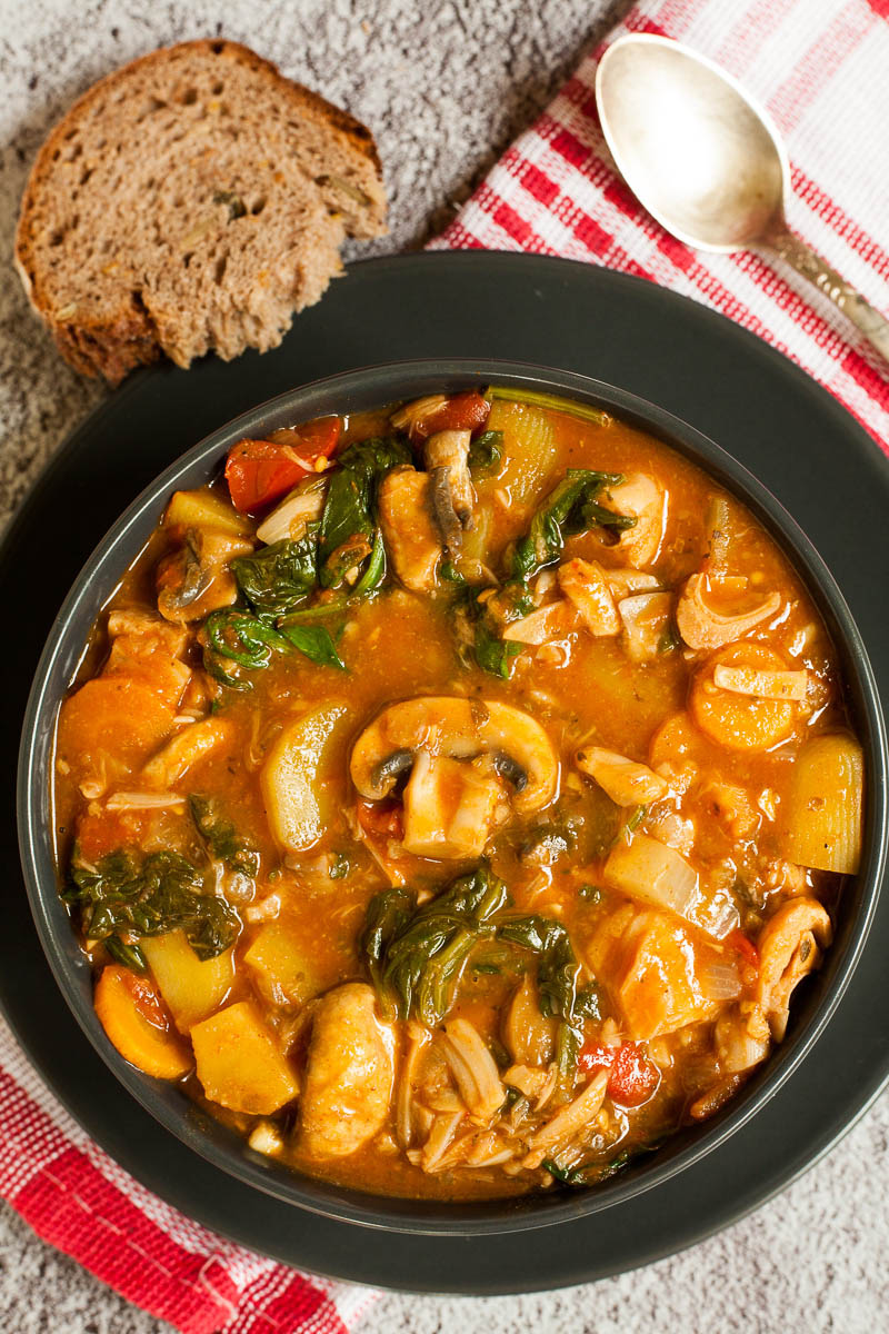 A black bowl from above with a stew where you can see chopped potatoes, spinach leaves, carrot slices, tomatoes, and mushroom slices. Slices of bread is next to it.