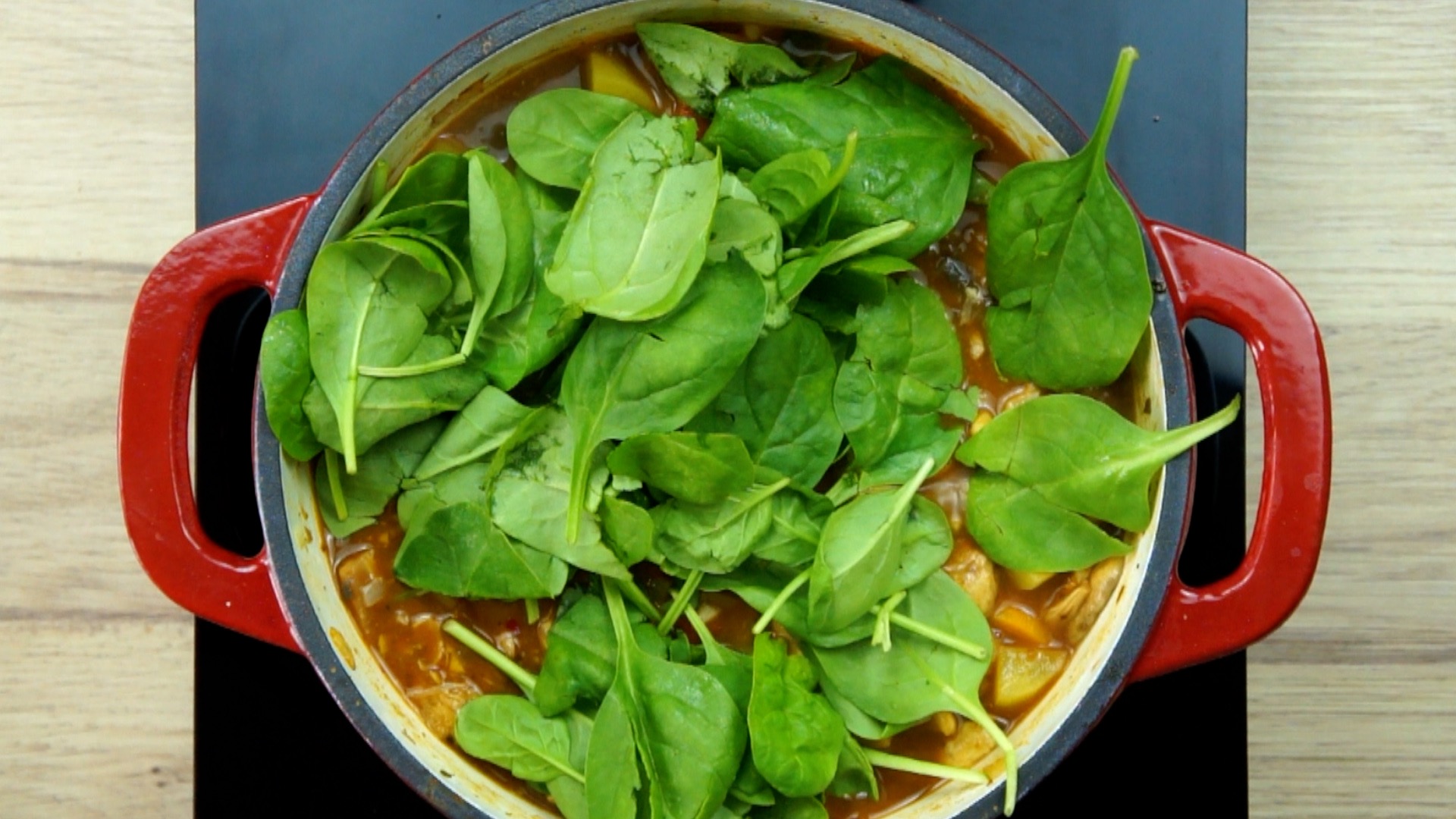 Red white Dutch oven with brown stew topped with fresh spinach leaves. 