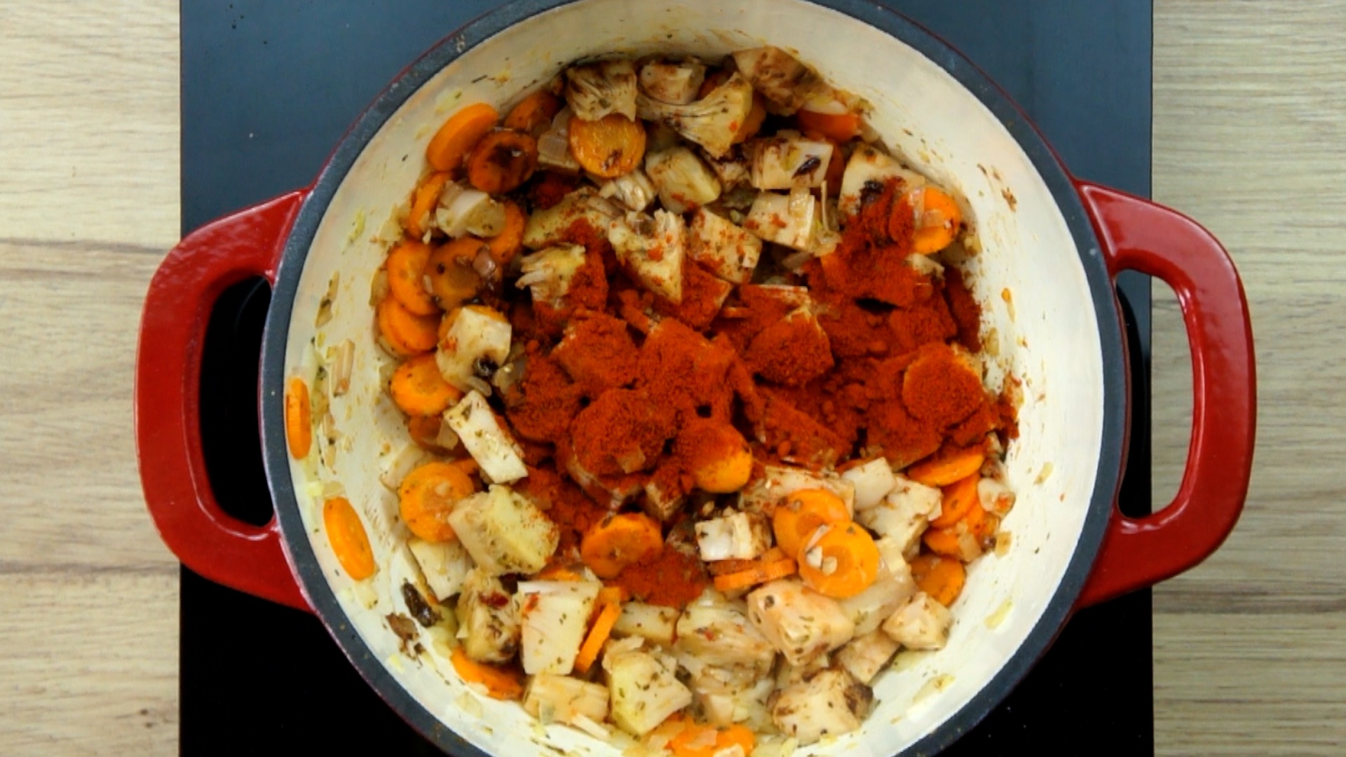Red white Dutch oven with chopped onion, garlic, sliced carrots and jackfruit chunks sprinkled with red pepper powder. 