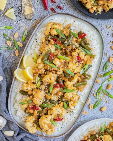 Large oval blue plate with white rice light brown sticky cauliflower, green beans, red chili peppers, scallion and black sesame seeds. Other portions are in side dishes and many ingredients are scattered around.