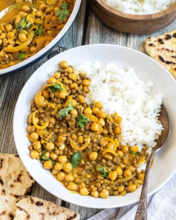 A white plate with white rice, chickpeas, and lentils, in a creamy light brown sauce sprinkled freshly chopped cilantro. A spoon is placed inside. Naan bread, a bowl of rice, and the remaining curry in the pan are next to it.