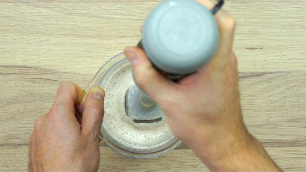A hand blender is placed in a tall glass container with a white liquid. 