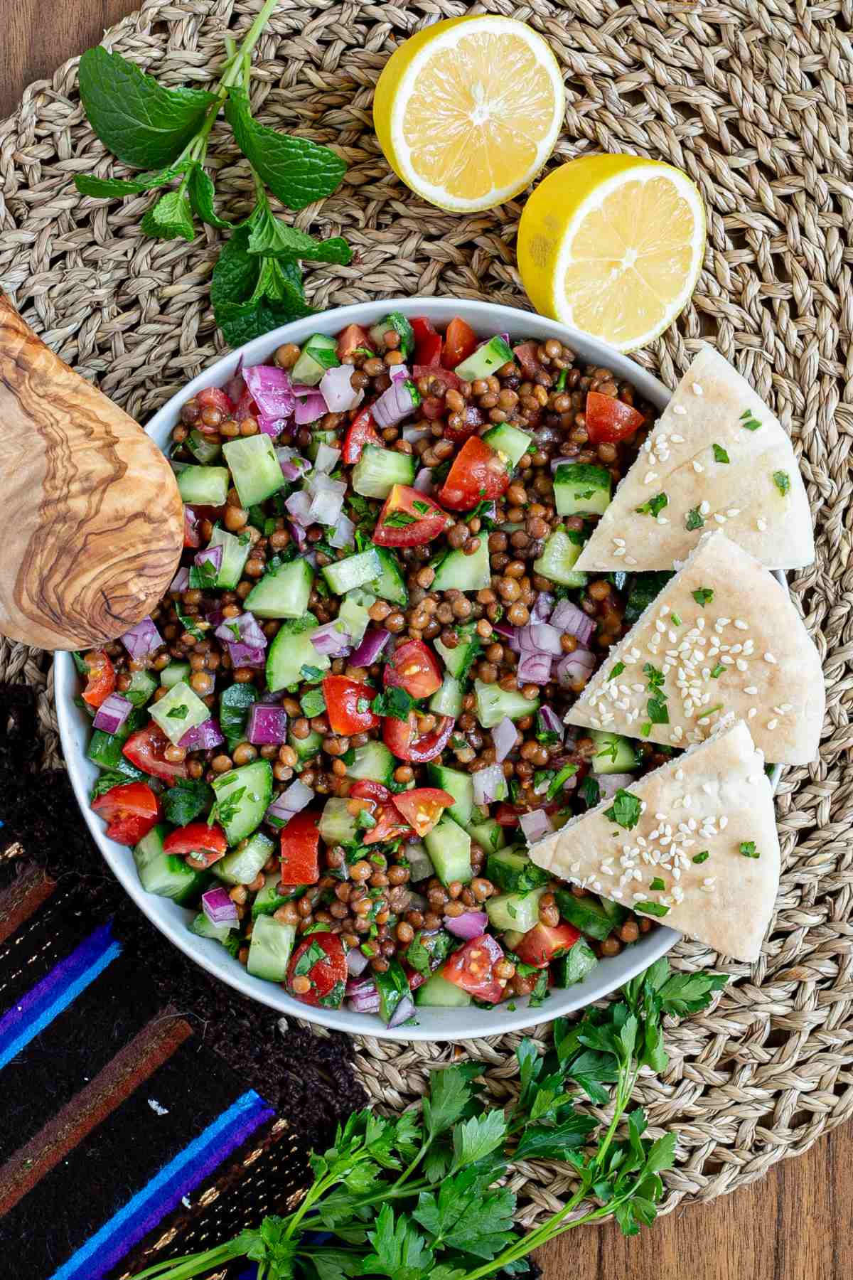 Large white bowl with a chopped mixed salad of brown lentils, cucumber, tomatoes, and red onion. 3 pita triangles are placed on the side.