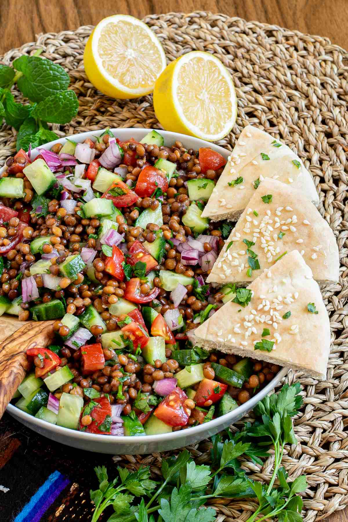 Large white bowl with a chopped mixed salad of brown lentils, cucumber, tomatoes, and red onion. 3 pita triangles are placed on the side.