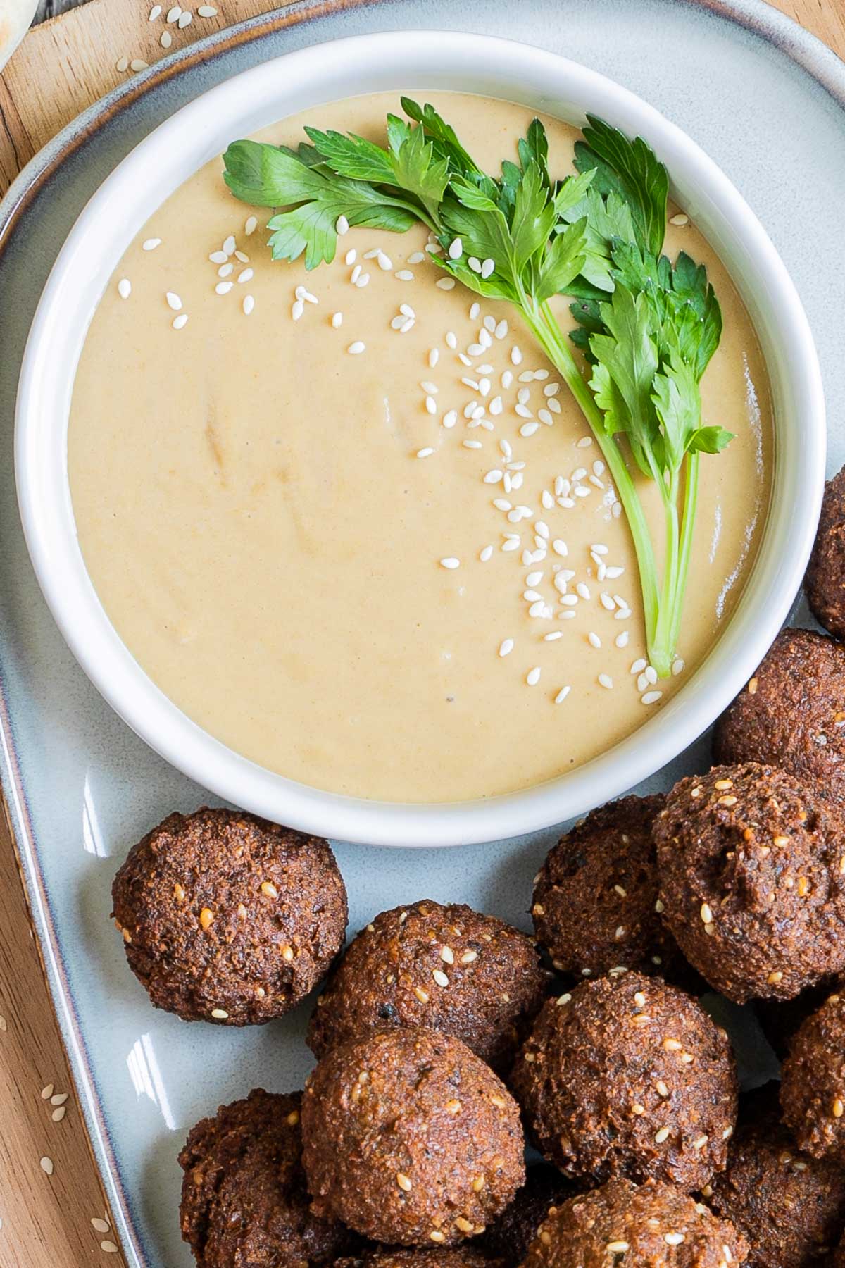Light brown sauce in a white shallow bowl sprinkled with white sesame seeds and topped with a twig of parsley. Falafel balls are next to it. 