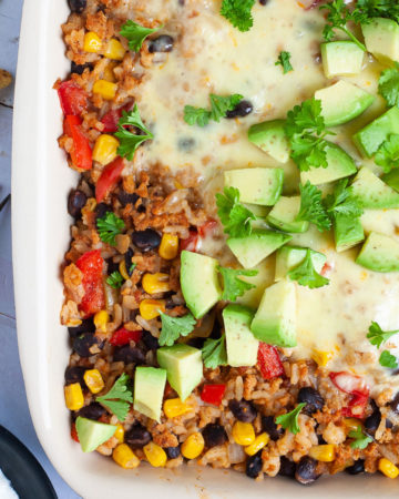A ligth brown ceramic dish with colorful ingredients like black beans, corn, rice, melted cheese, bell pepper strips, chopped avocado, chopped green herbs.