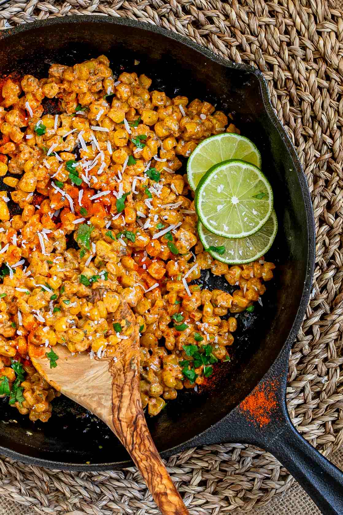 Cast iron skillet with charred yellow corn covered in spices and chopped herbs. A wooden spatula and two lime slices are placed inside.