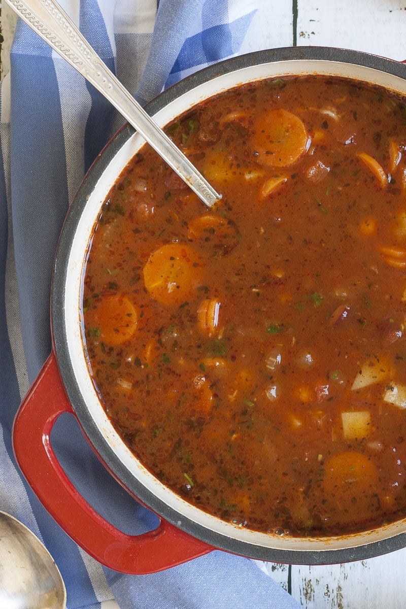 Red soup in a white dutch oven full of carrot slices, diced potatoes, beans, chickpeas and chopped green herbs. 