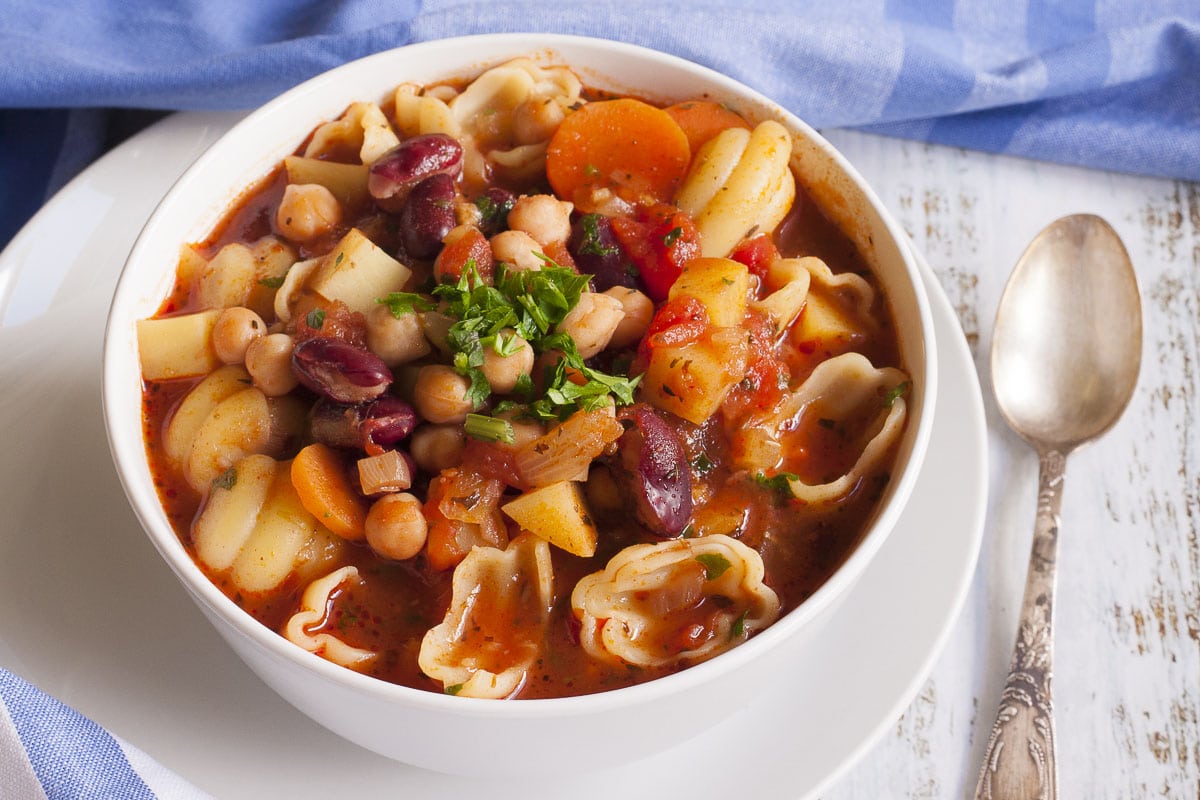A white bowl with a red soup full of red kidney beans, chickpeas, potatoes, carrots and shell pasta. It is topped with fresh parsley. 