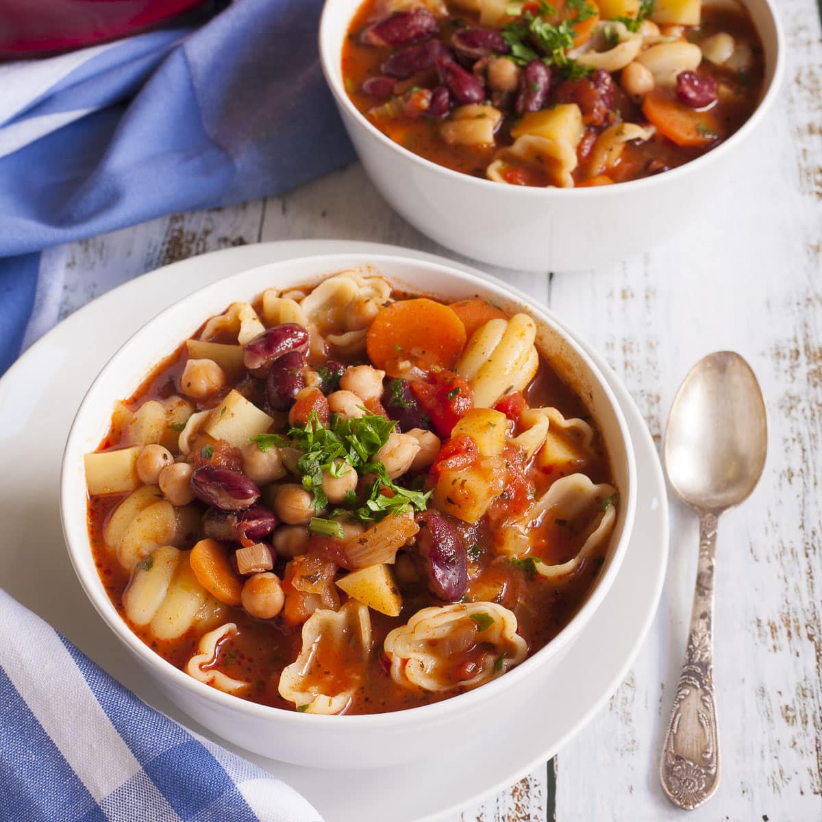 2 white bowls with a red soup full of red kidney beans, chickpeas, potatoes, carrots and shell pasta. It is topped with fresh parsley. 