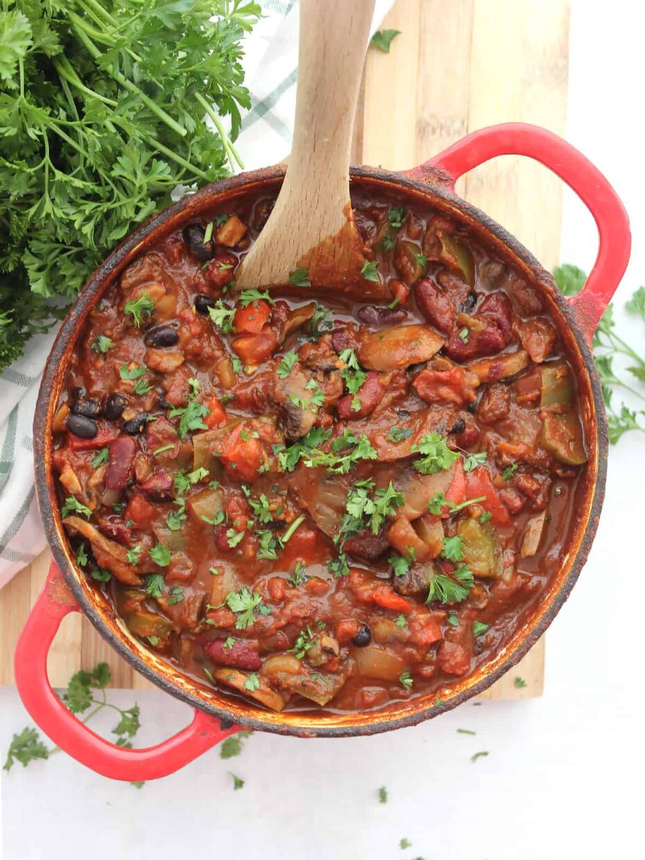 A red Dutch oven from above full of chili so red sauce, black beans, green hot pepper slices, shredded cheese, mushroom slices, chopped cilantro