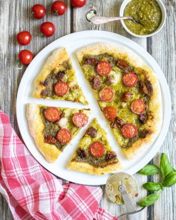 Sliced pizza on a white pizza plate topped with green pesto, melted cheese, sun-dried tomato pieces, and halved cherry tomatoes. More pesto and fresh ingredients next to the plate.