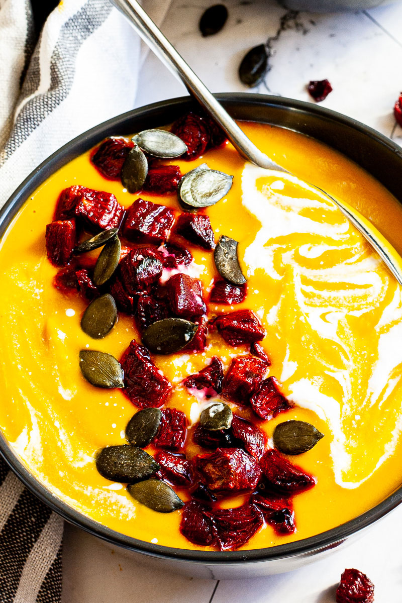 Yellow cream soup in a black bowl topped with diced roasted beetroot, green pumpkin seeds and drizzled with a white sauce.