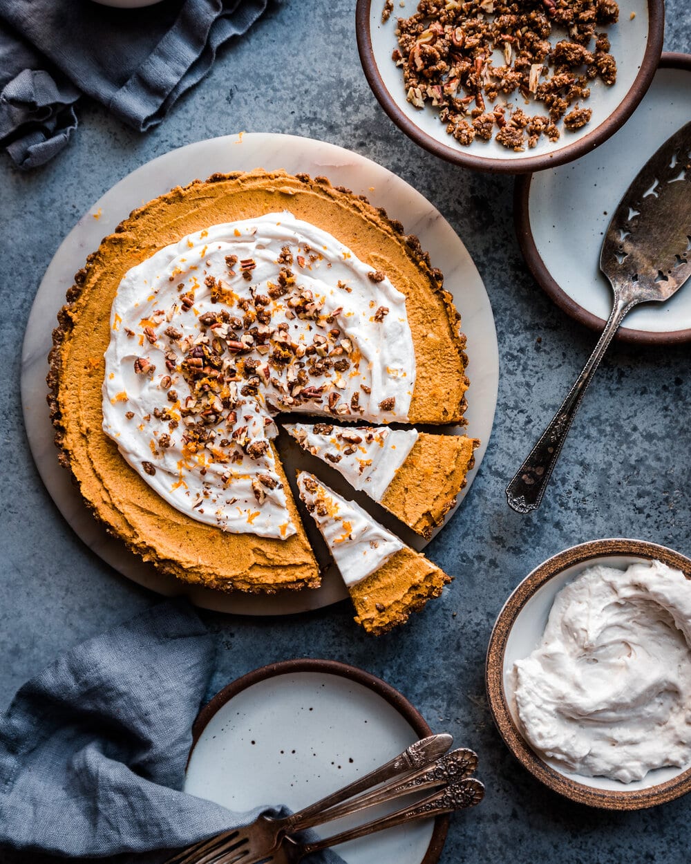 Pumpkin pie with whipped cream and pecans next to smaller plates with chopped pecans and whipped cream.