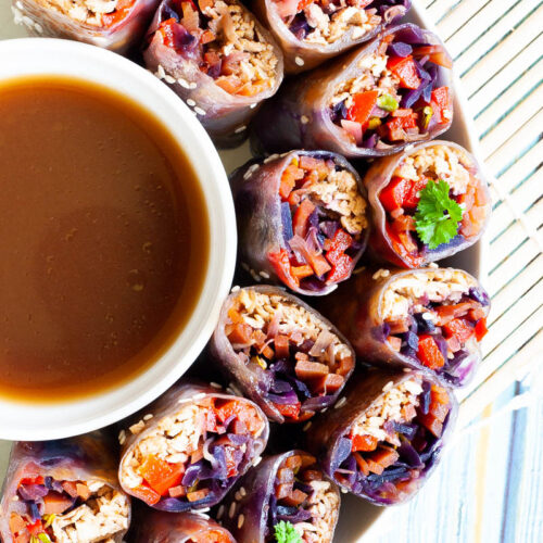 Lots of spring rolls cut in half and arranged in a semi-circular dish facing upwards around a brown dipping sauce.