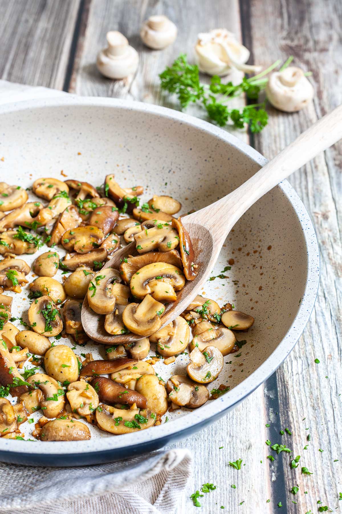 Golden brown sliced button and shiitake mushrooms in a frying pan topped with freshly chopped greens. A wooden spoon is taking some from the middle.