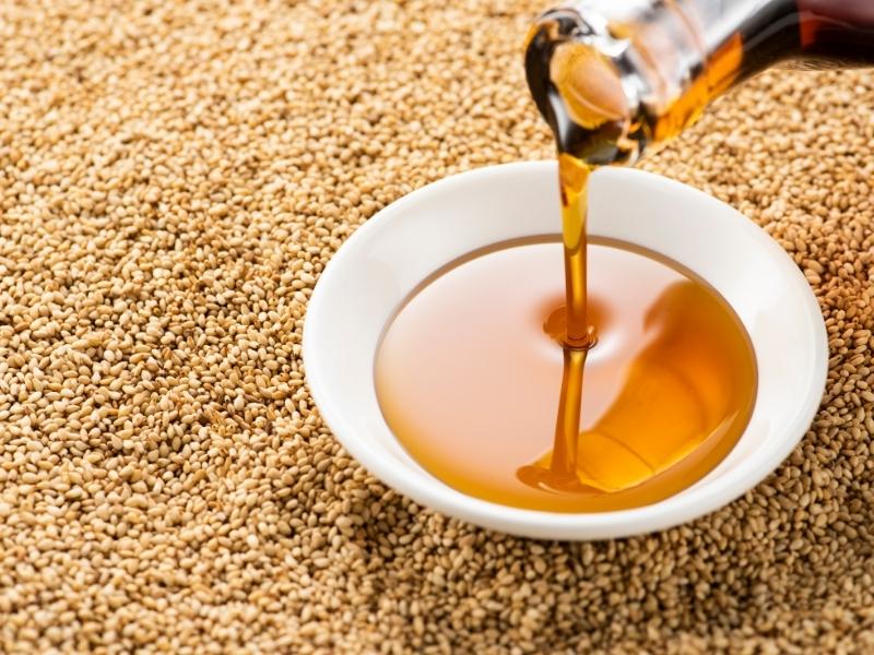 A small white plate is on top of sesame seeds. Oil is pouring out of a glass bottle on the plate. 