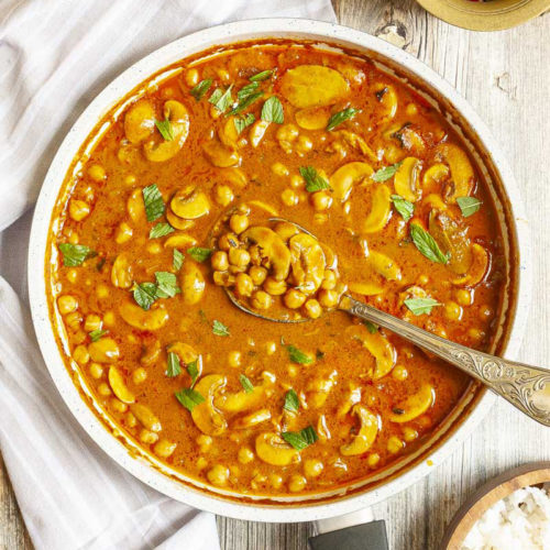 Large saucepan from above with brown-yellow liquid chickpeas and mushroom slices sprinkled with chopped green leaves