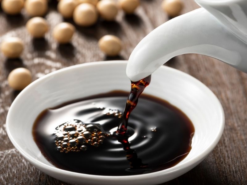 Dark brown liquid is poured into a small white bowl. Soybeans are behind the bowl. 