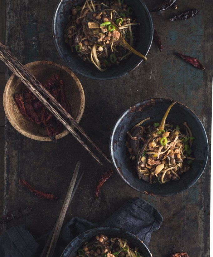 An overhead image of several small bowls of eggplant stir-fry on a rustic metal tabletop.
