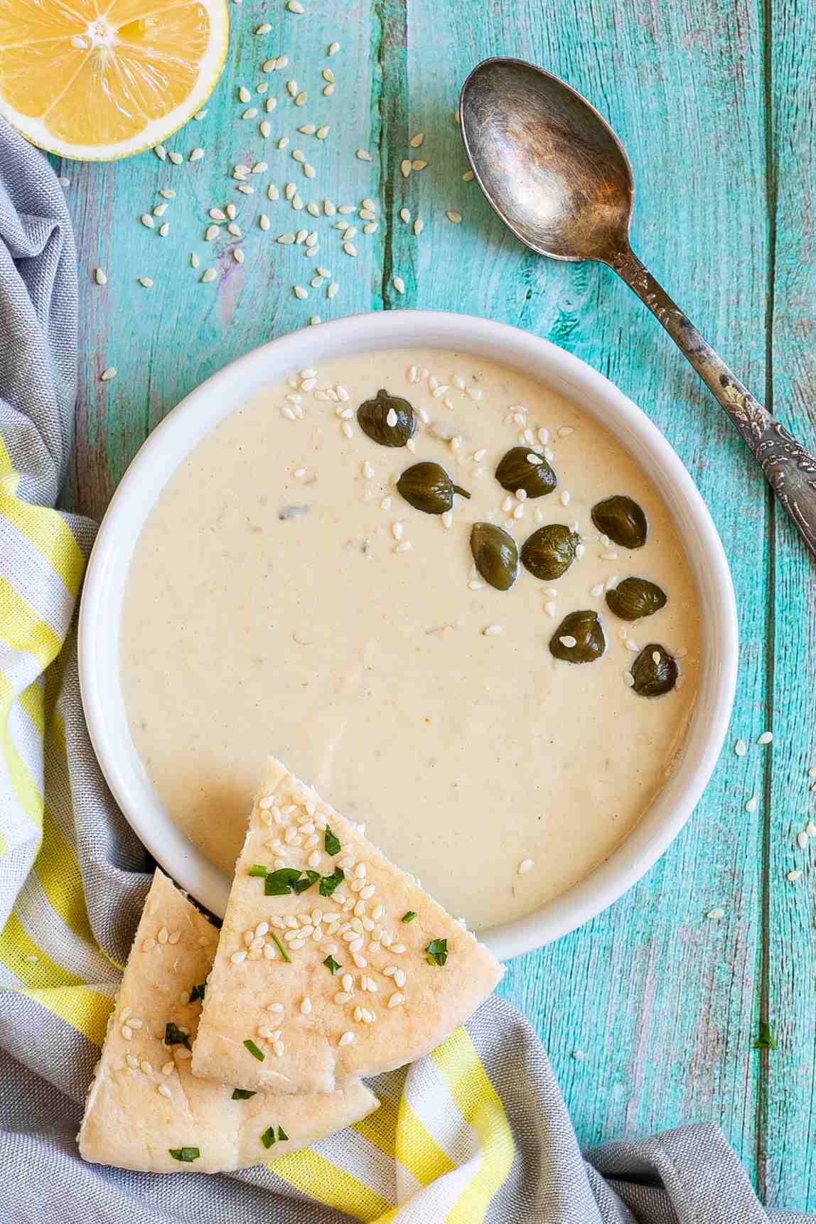 Small white bowl with a light brown thick sauce topped with sesame seeds and capers. Two pita triangles are placed on the side.