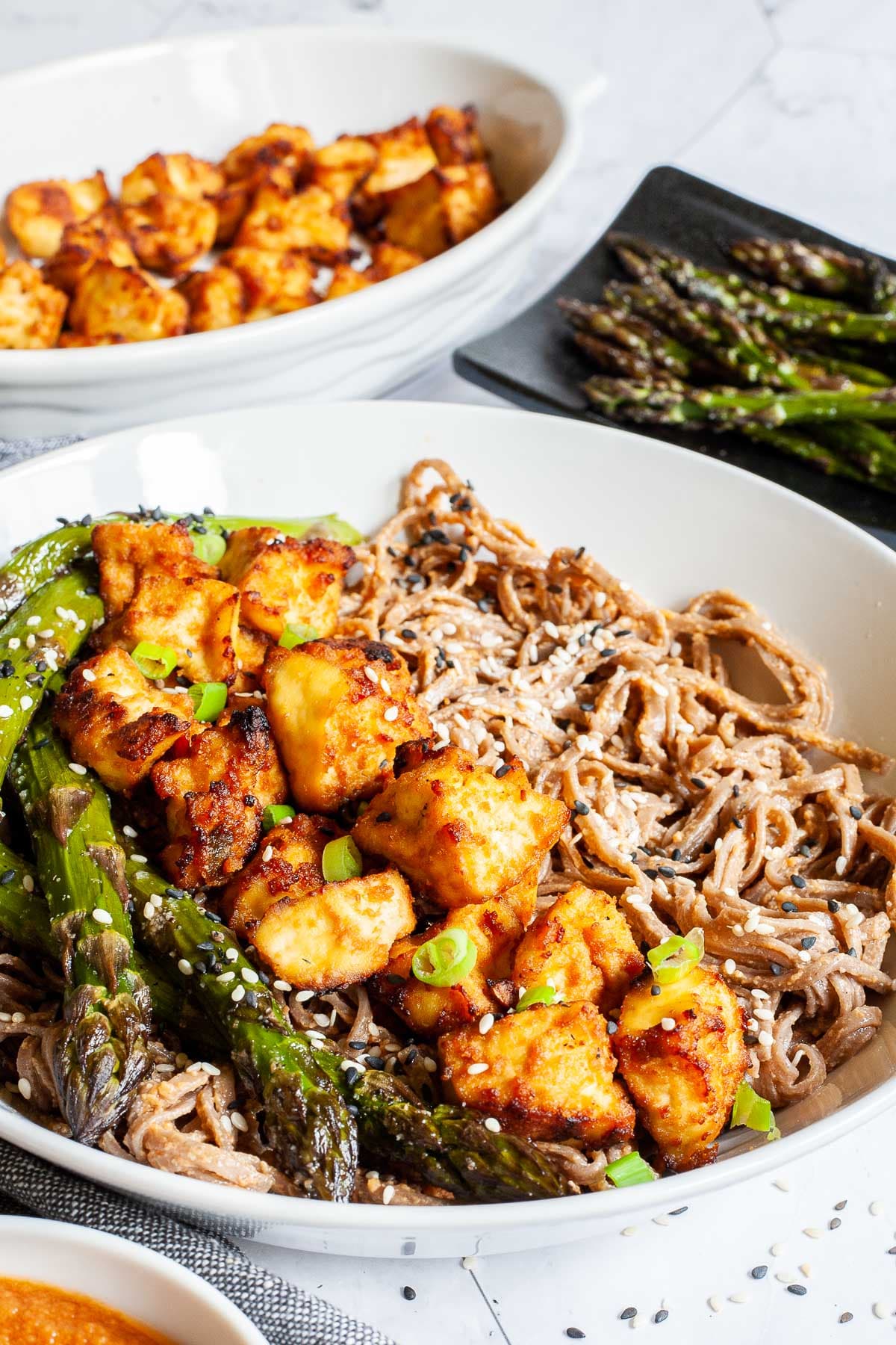 Large white bowl with brown soba noodles, orange crispy tofu bites, roasted asparagus and black and white sesame seeds. More asparagus and tofu bites are on the side.