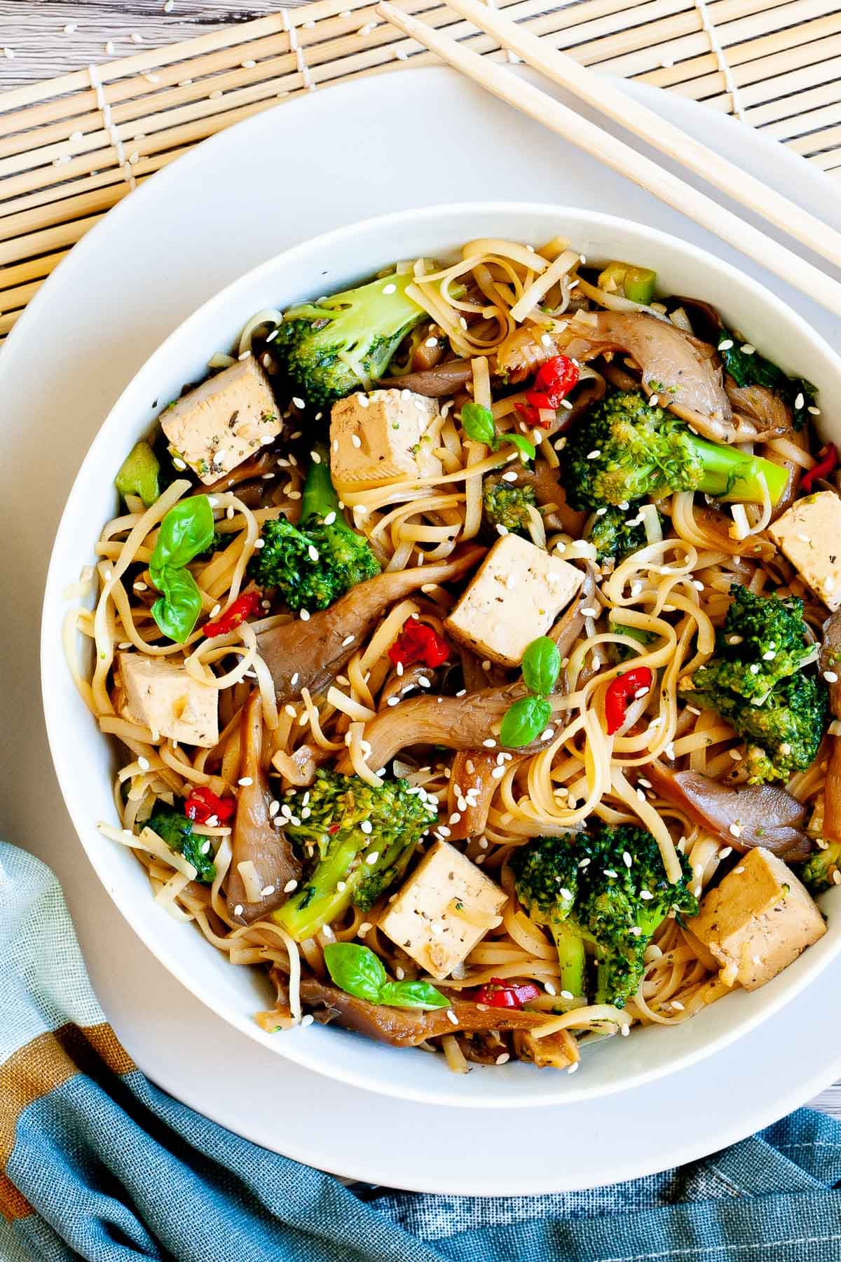 White bowl with tofu cubes, broccoli florets, oyster mushroom shreds, red chili slices and long thin rice noodles.