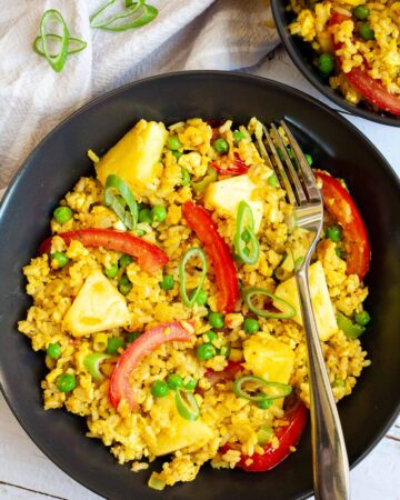 2 black bowls with yellow rice, red tomato slices, pineapple chunks, green peas and chopped green onion.