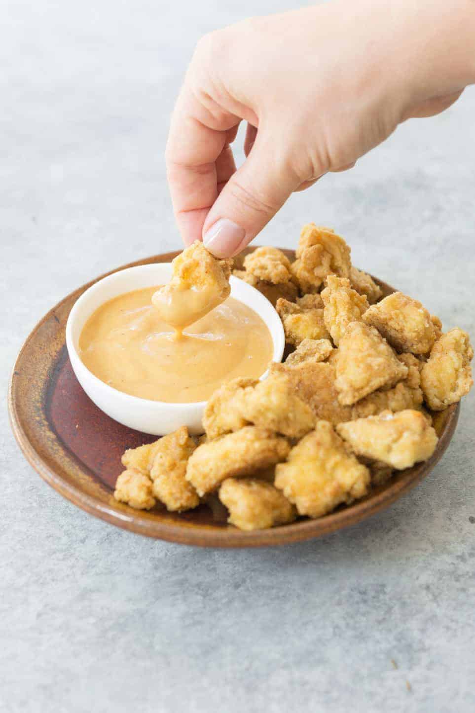 Several small light brown nuggets on a brown plate. A hand is dipping one into a pink-orange sauce in a small bowl right next to them.
