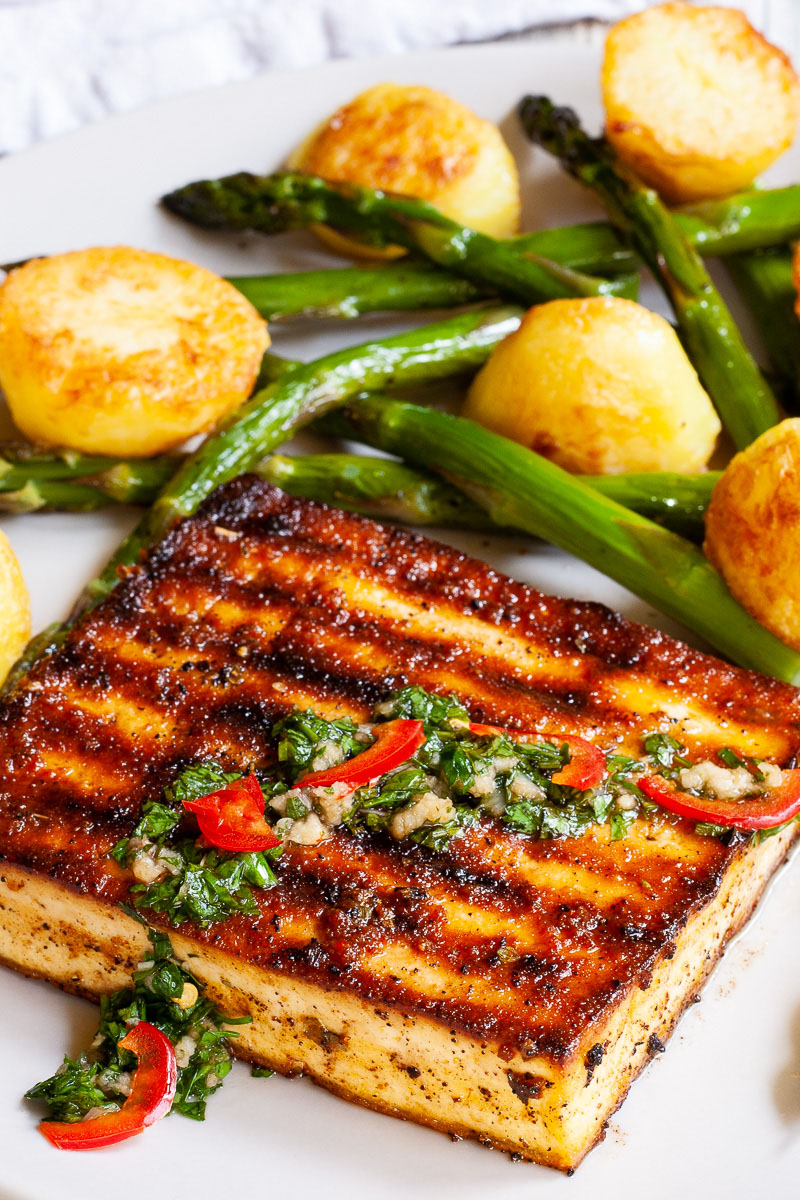 Brown, shiny tofu slice up close topped with a chopped green herbs served with baked half potatoes and roasted asparagus in the background