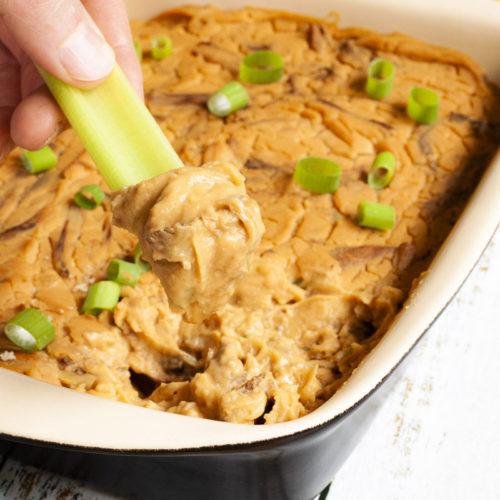 A hand is holding a green celery stick that was dipped in an orange dip with brown shreds. The dip is at the background in a light brown square dish.