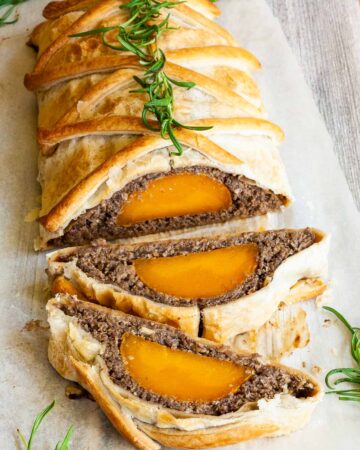 2 slices of vegan wellington on the side. Yellow squash with brown puree around wrapped in a pastry shell. Green herbs are around.