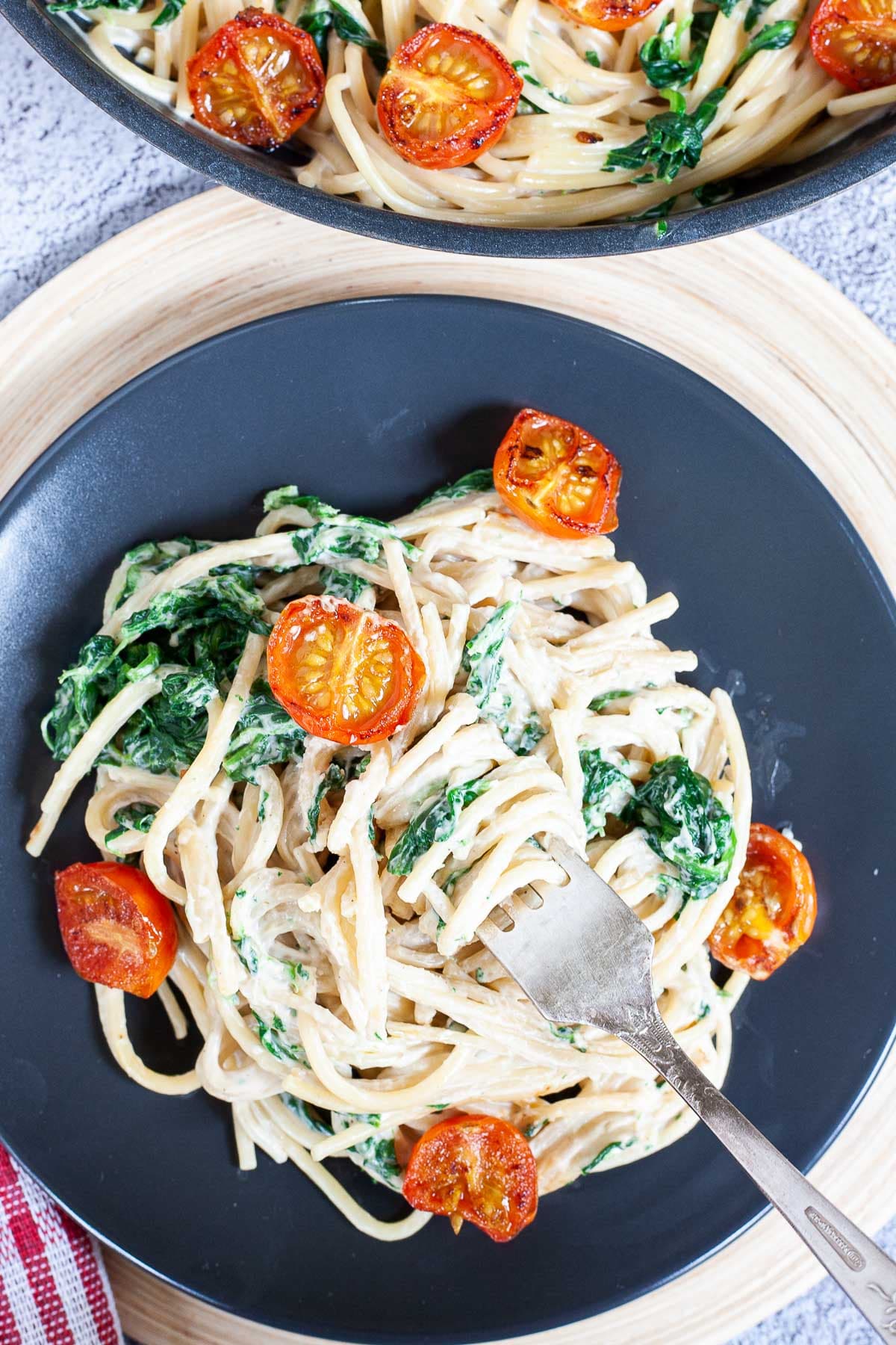 Spaghetti in white sauce is served on a black plate with charred cherry tomatoes and wilted spinach leaves. The remaining pasta is in a black frying pan next to it.